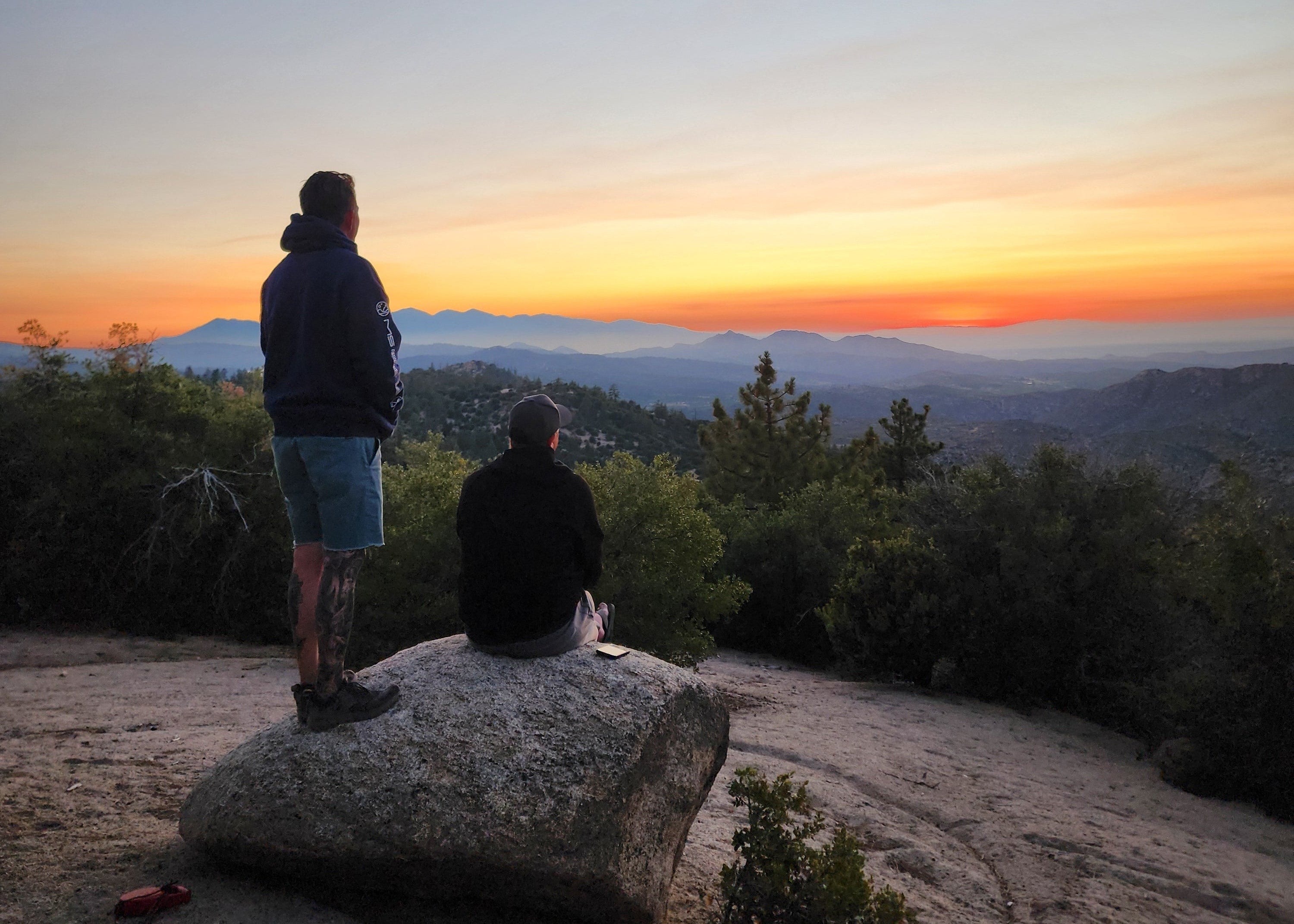 Two people standing on a rocky outcrop with a colorful sunset or sunrise in the background.